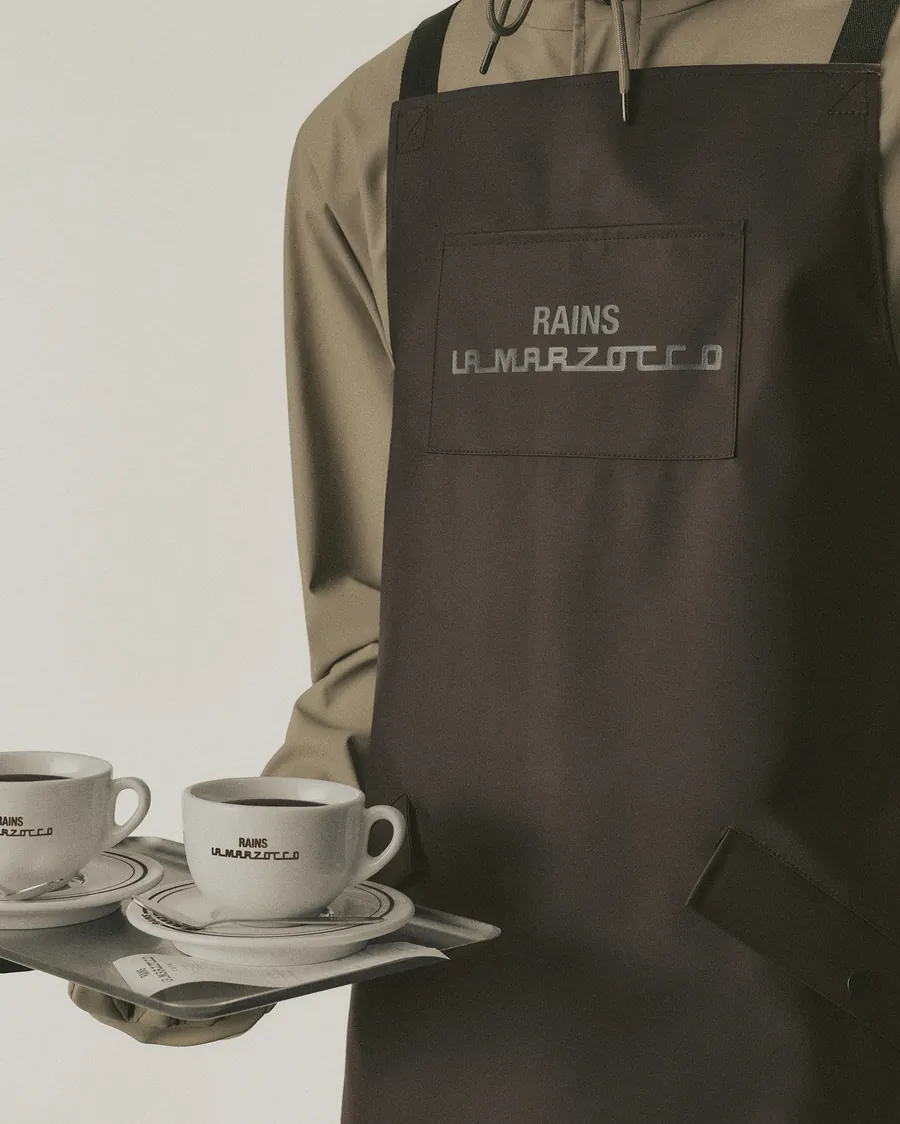 Delantal de barista marrón de Rains y La Marzocco con el logotipo en el bolsillo del pecho y una bandeja con dos tazas