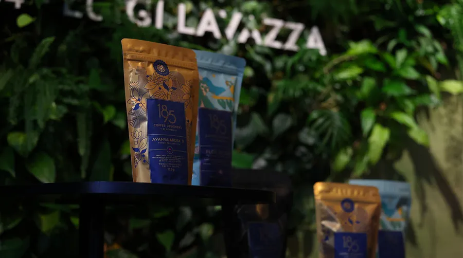 Paquetes de café de 1895 Coffee Designers by Lavazza delante de una pared vegetal con el rótulo de Lavazza